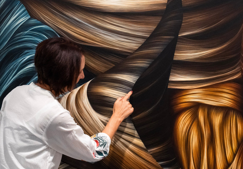 A woman in a white shirt and patterned skirt points at a large abstract wall painting with flowing brown, black, gold, and blue brushstrokes.